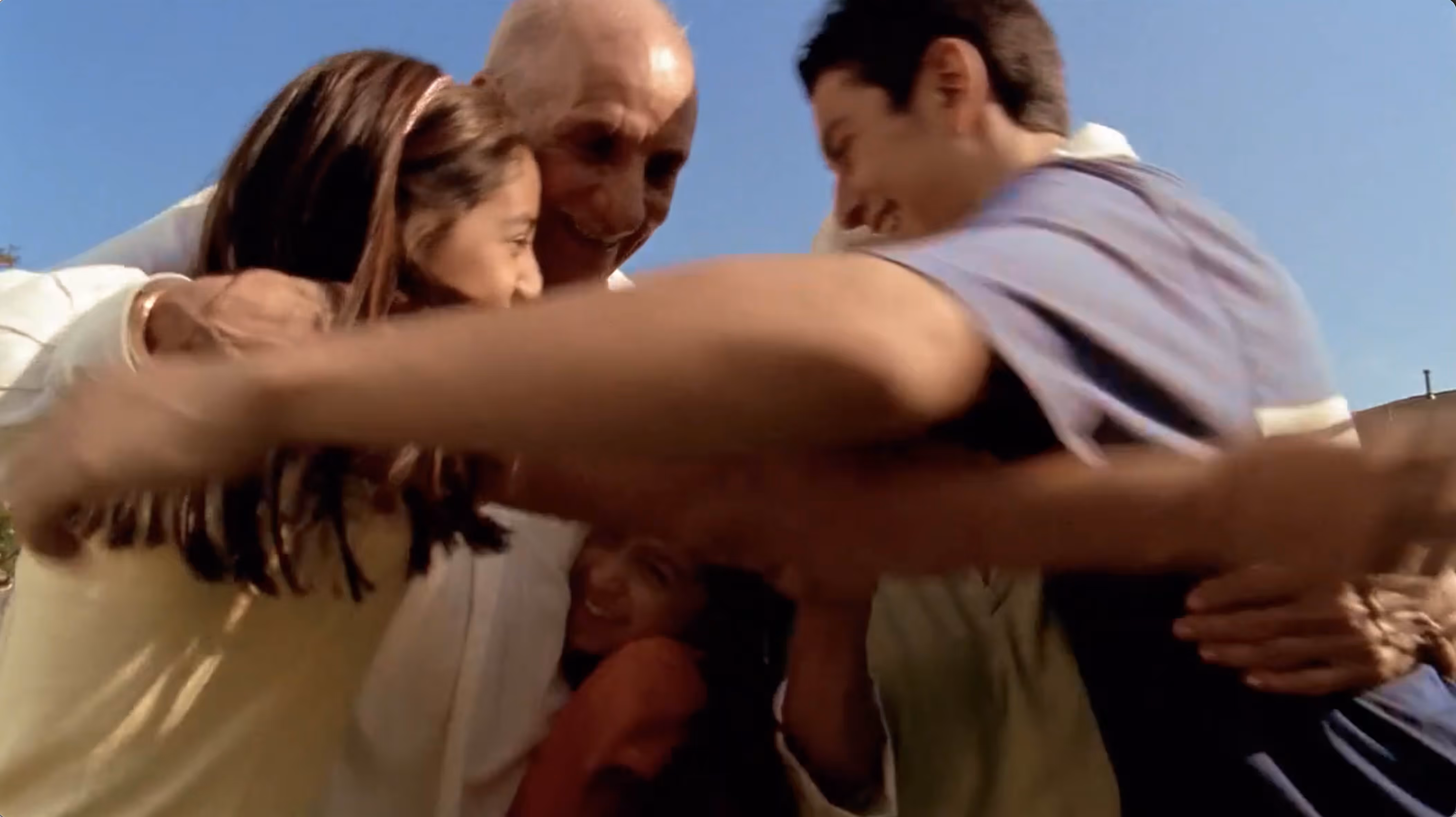 A group hug of smiling people outdoors under clear blue sky, showing affection and togetherness.