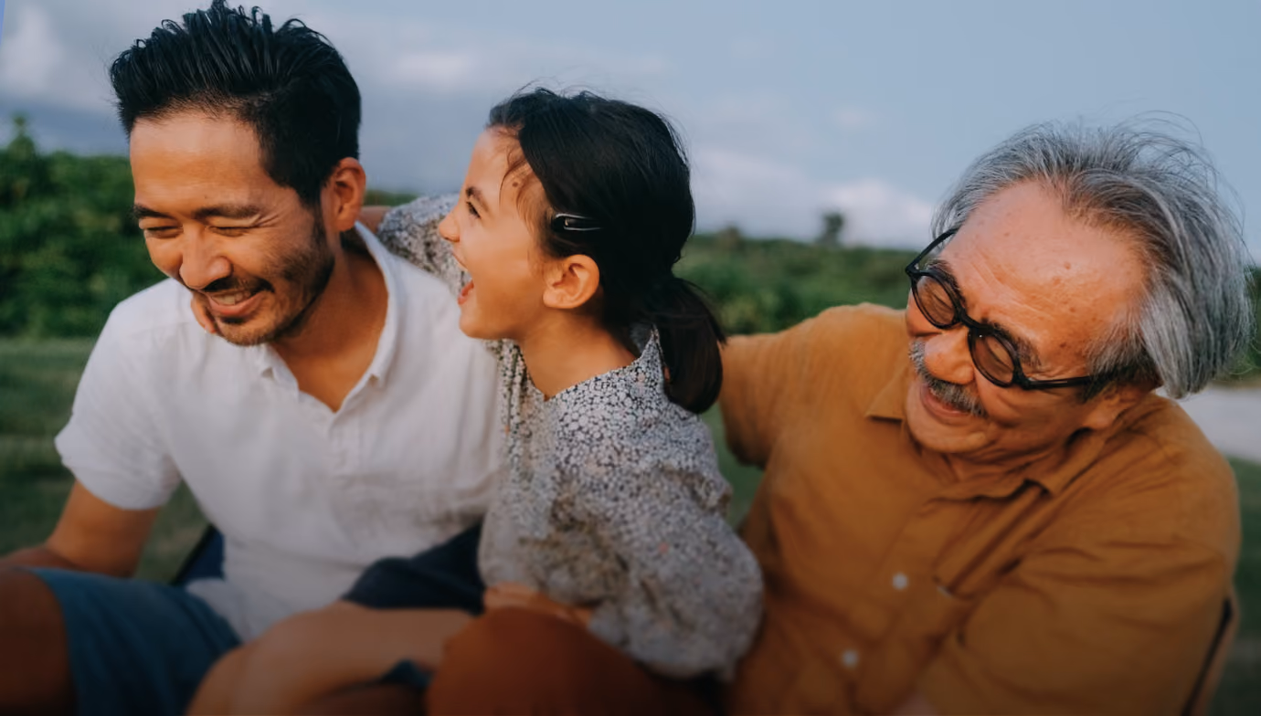 Three generations laughing outdoors: a young girl between two men, one elderly and one middle-aged.
