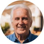 Smiling elderly man with white hair wearing a blue collared shirt outdoors.