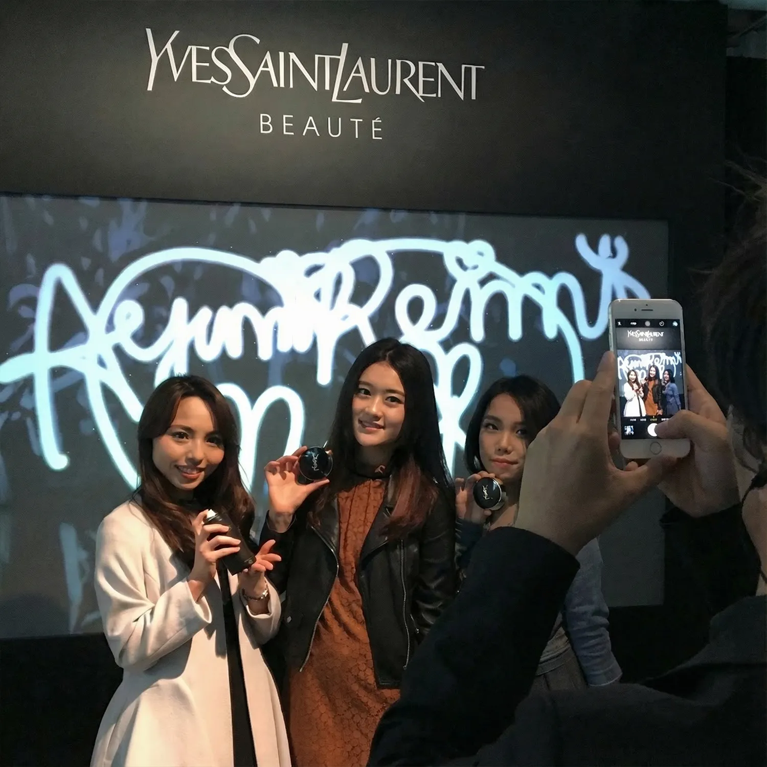 Three women posing with Yves Saint Laurent beauty products while a person takes their photo with a smartphone in front of a branded backdrop.