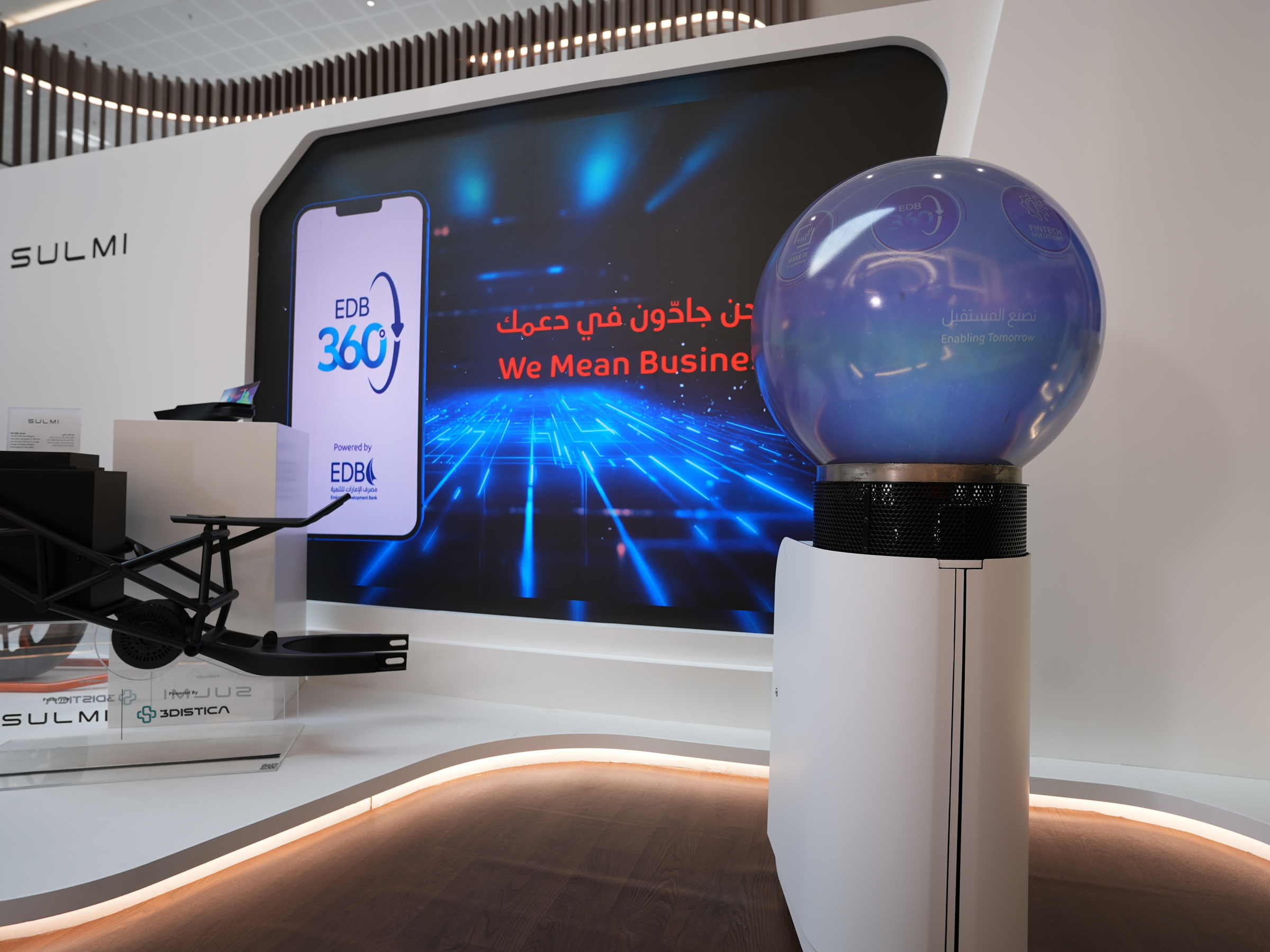Futuristic tech display featuring a large globe-shaped device with 'Enabling Tomorrow' text, a digital screen with EDB 360° app promotion, and a black mechanical frame labeled SULMI.