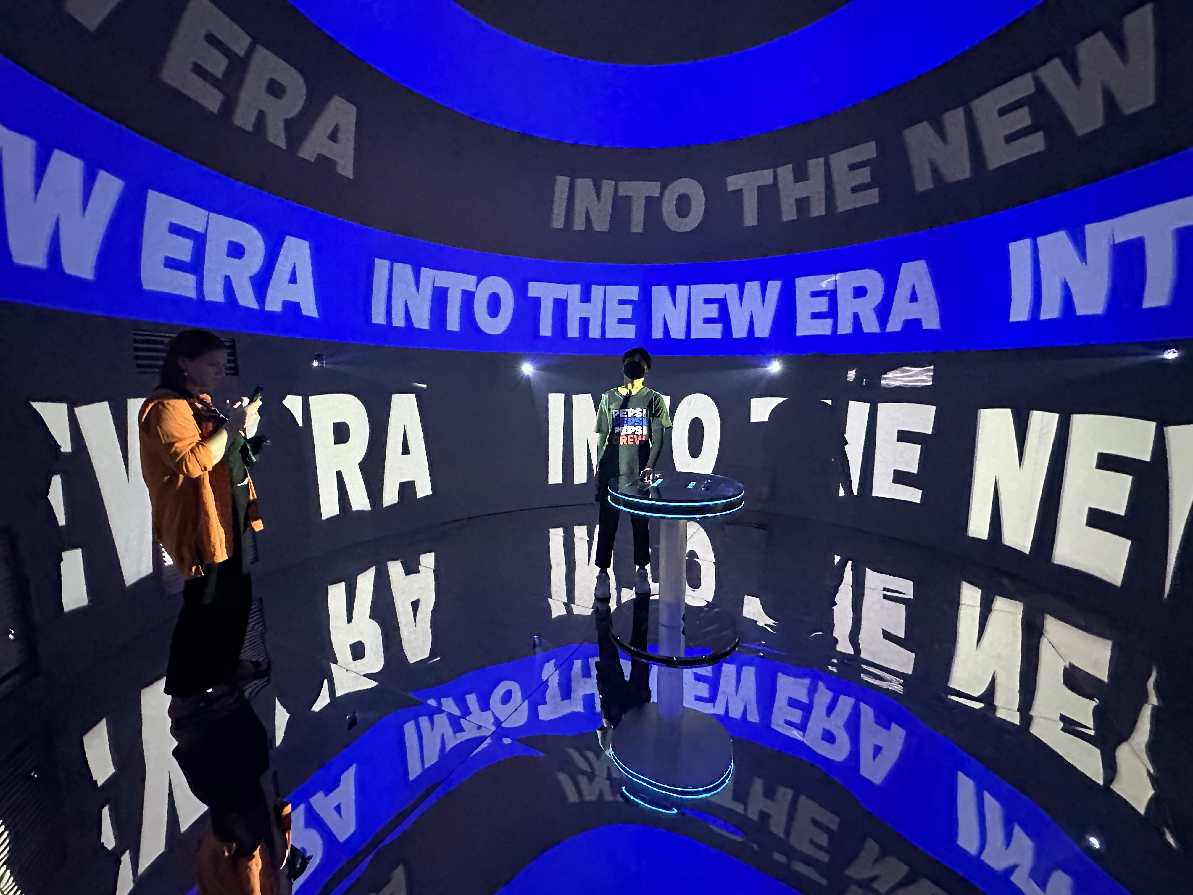 Man standing at a circular table in a dark room with reflective floor, surrounded by projected text reading 'INTO THE NEW ERA'.