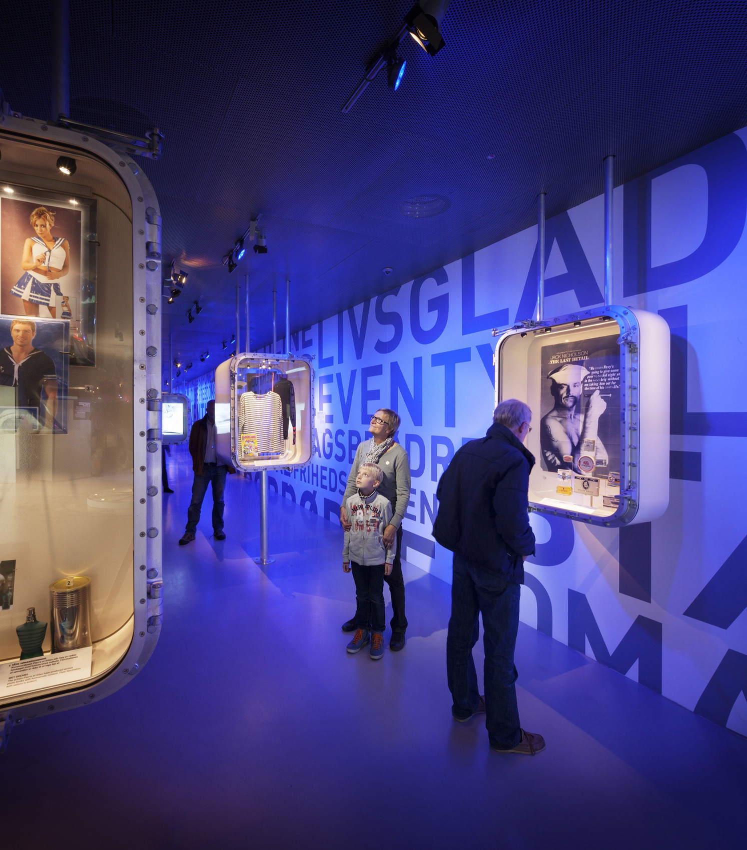 Museum exhibition with people viewing illuminated display cases mounted on poles against blue walls with large letters.