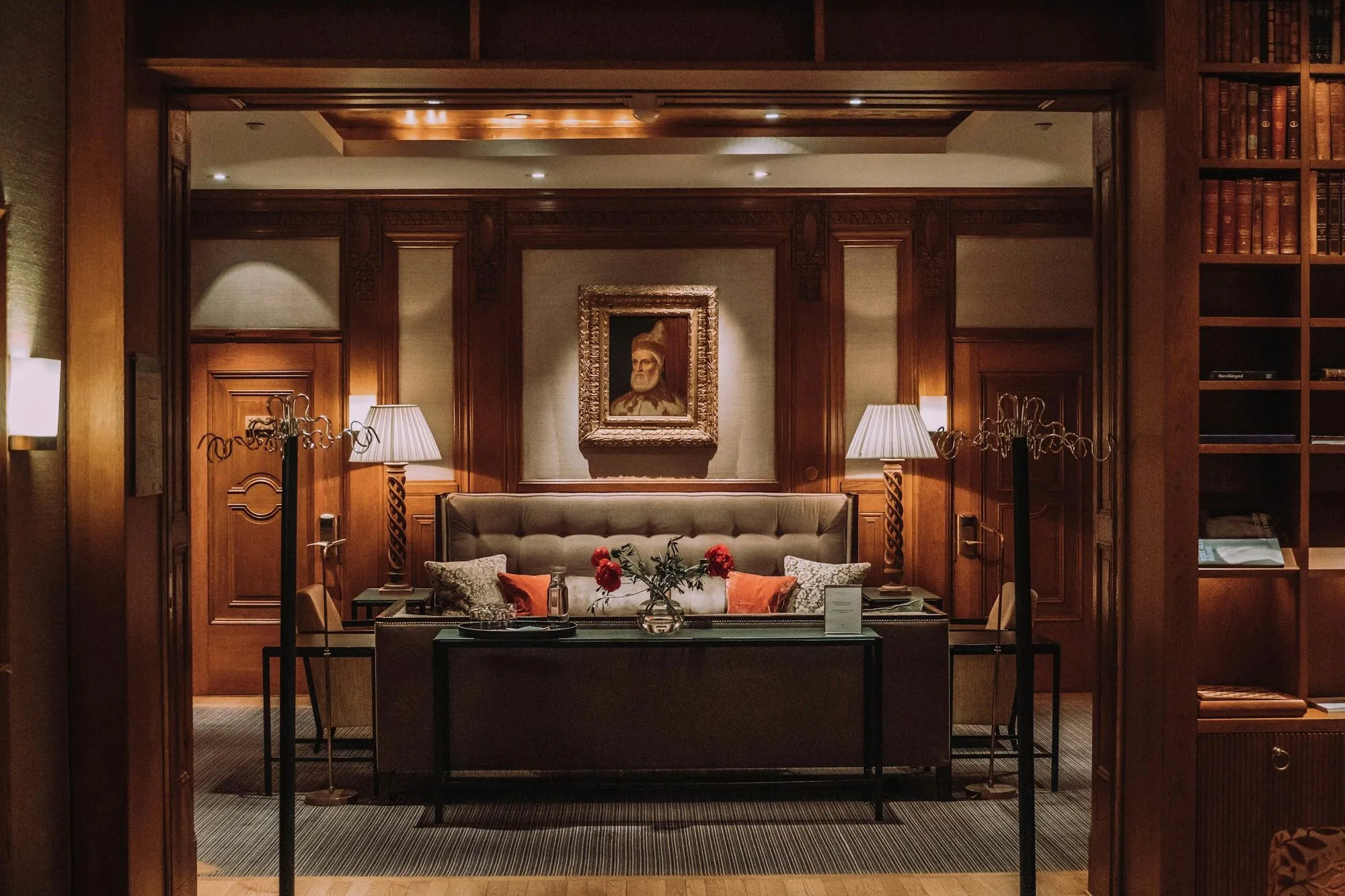 Cozy, warmly lit sitting room with a tufted sofa, patterned and orange pillows, a glass vase with red flowers on a table, wooden paneled walls, two lamps, a framed portrait, and bookshelves.