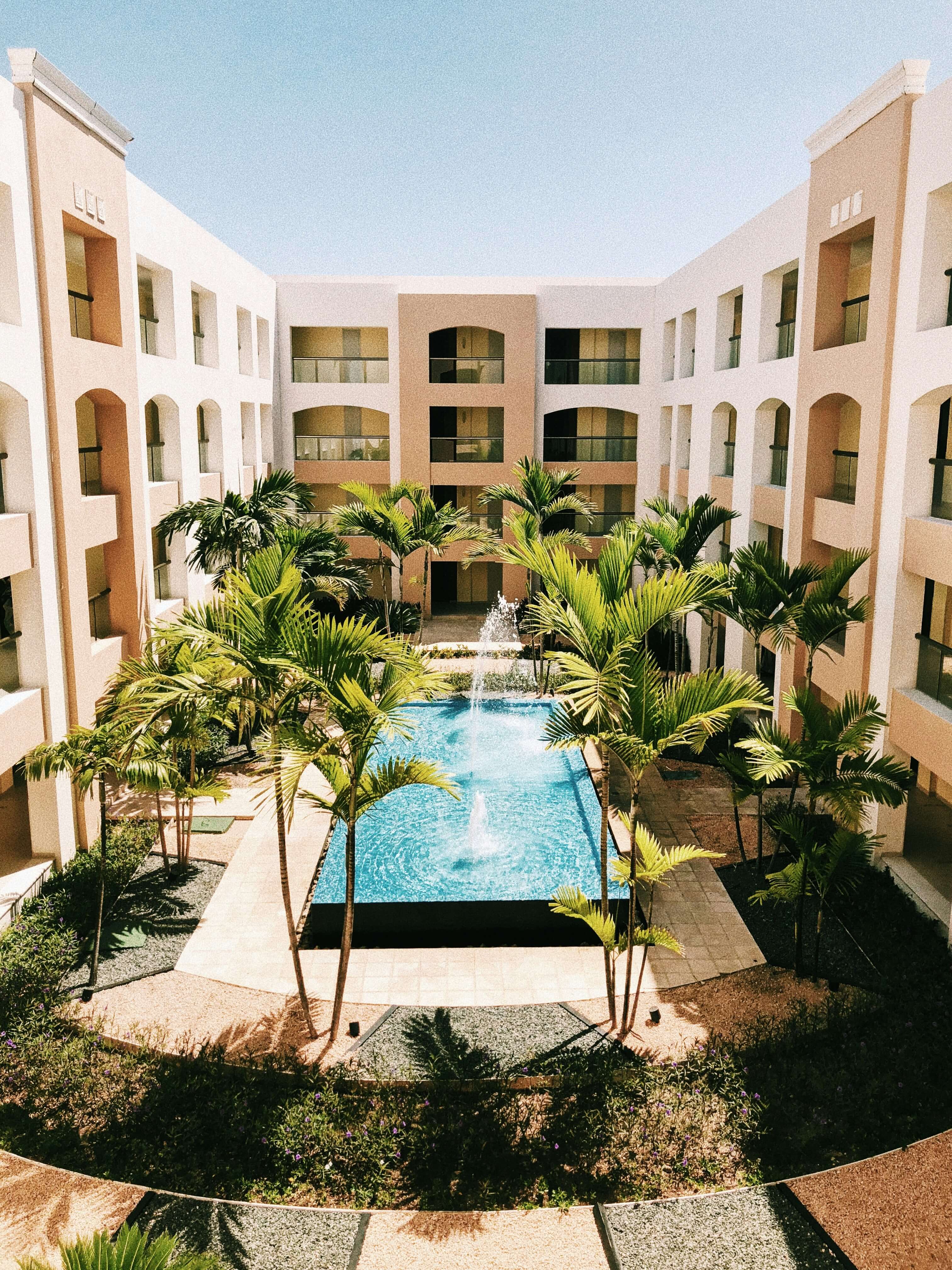 Courtyard of a modern apartment complex with a central fountain and palm trees surrounding a clear blue pool.