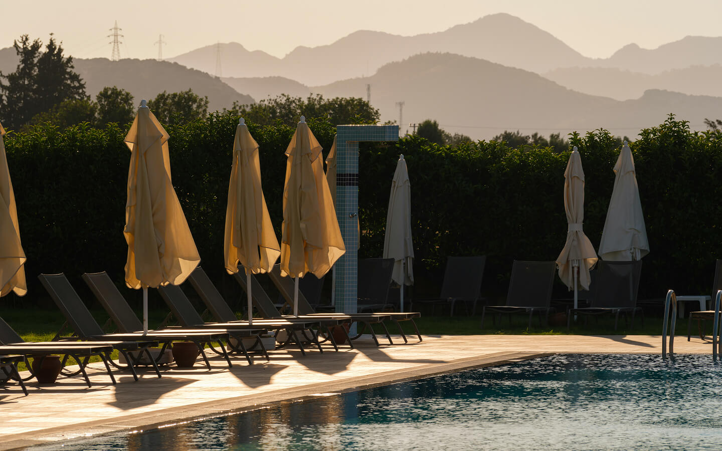 Empty poolside loungers with closed beige umbrellas and a tiled outdoor shower at sunset, with mountains in the background.
