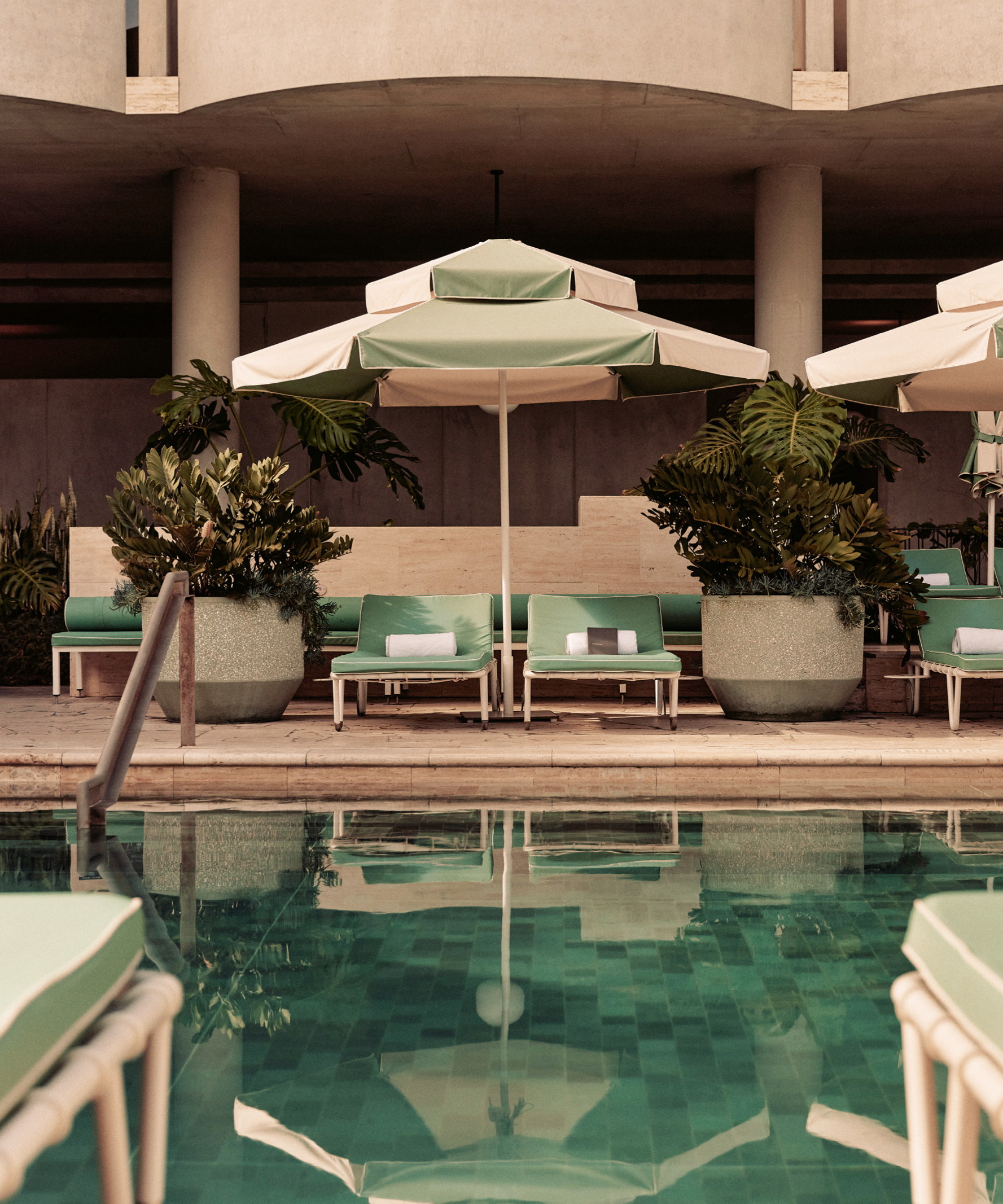 Green cushioned lounge chairs and a green and white umbrella by a pool with reflective water and large potted plants nearby.