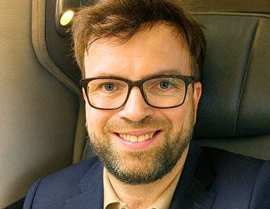 Smiling man with glasses and a beard sitting in a seat with dark upholstery.