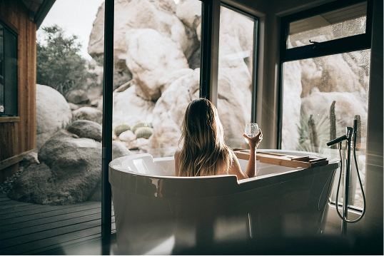 Woman with long hair relaxing in a modern bathtub holding a glass, with large windows showing rocky outdoor scenery.