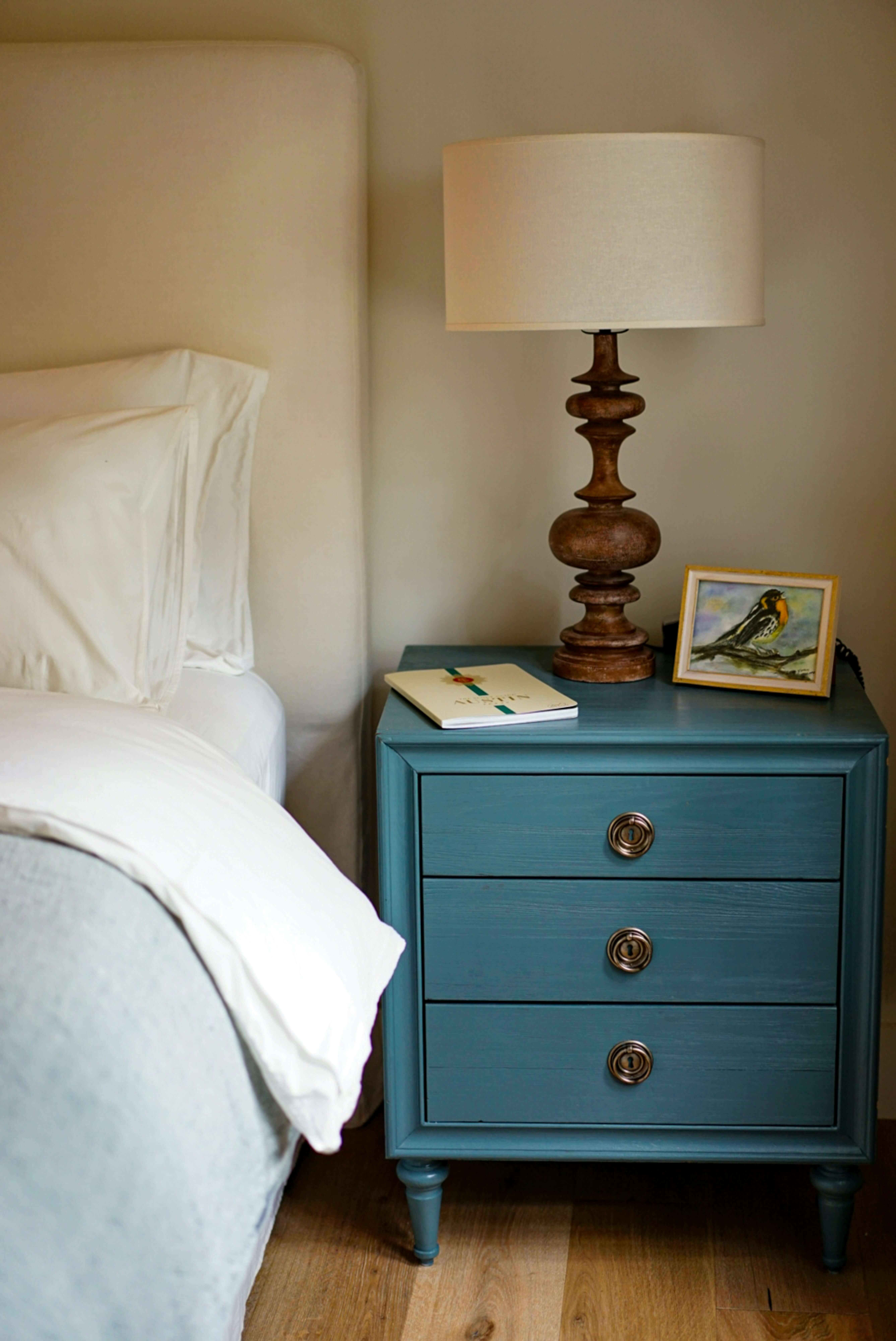 Blue wooden bedside table with three drawers, a decorative lamp, a book, and a framed bird painting next to a bed with white linens.