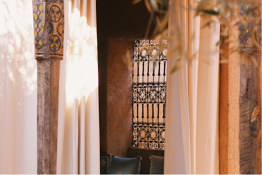 Cozy window with ornate wrought iron grille framed by soft beige curtains and patterned wooden pillar.