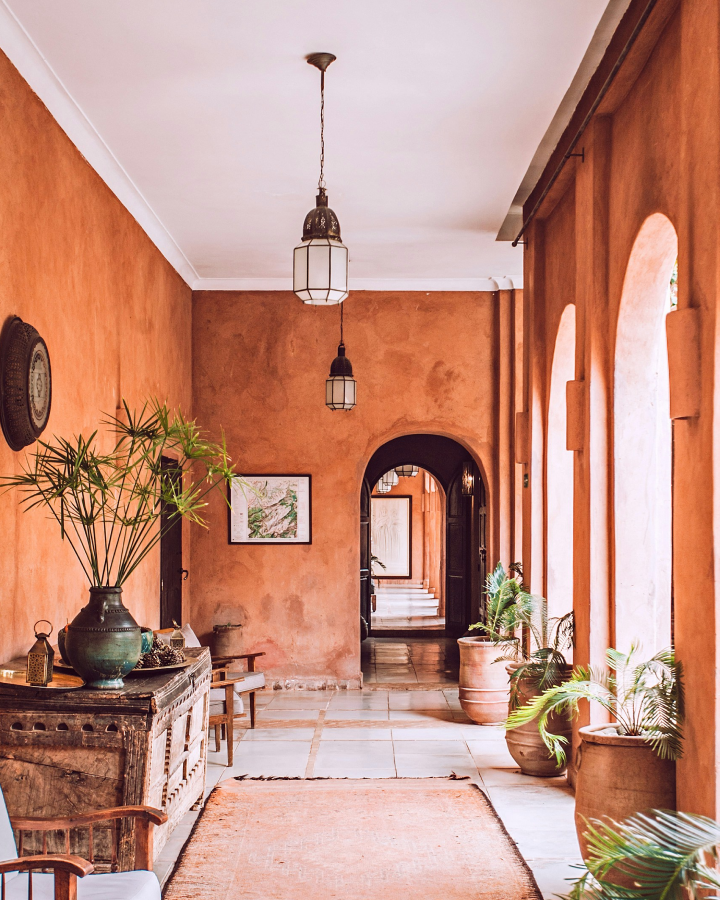 Rust-colored hallway with arched doorways, wooden furniture, hanging lanterns, and potted plants along the walls.