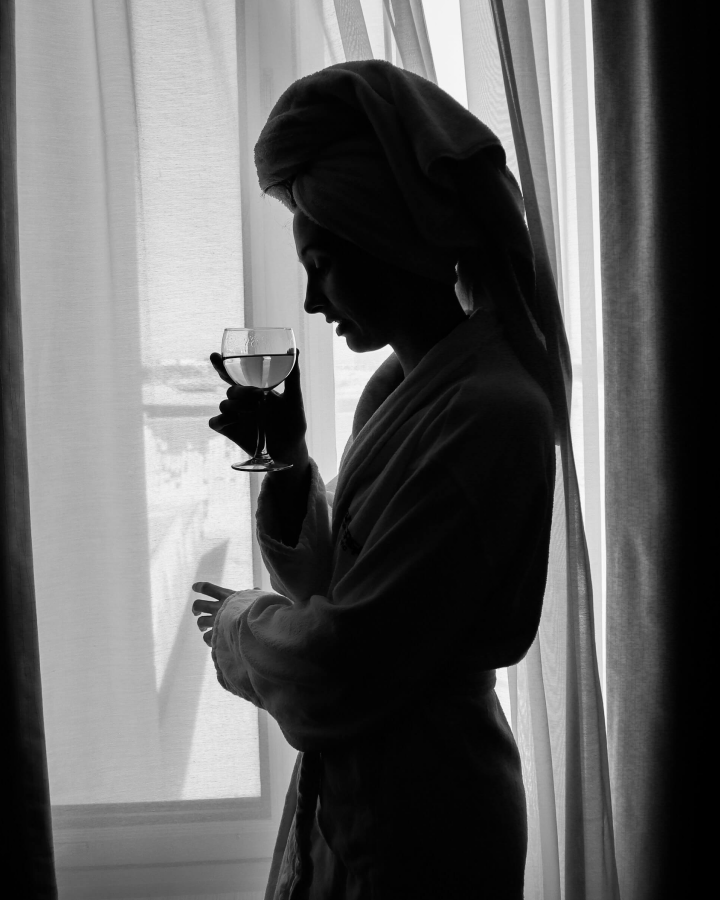 Silhouette of a woman in a bathrobe and towel on her head holding a wine glass near a window with sheer curtains.