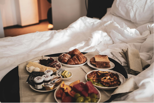 Breakfast tray on bed with croissants, toast, fruit, cheese, cold cuts, and small jars of spreads.