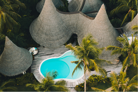 Aerial view of a tropical resort area with a uniquely shaped turquoise swimming pool surrounded by wooden decking and thatched-roof huts amidst palm trees.