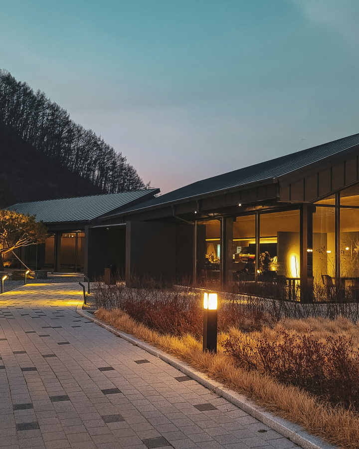 Evening view of a modern building with large windows and warm interior lighting, beside a paved walkway with lit lamps and dry bushes.