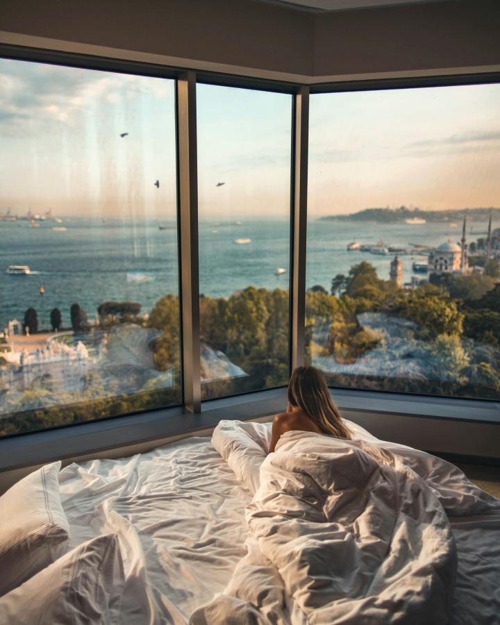Woman wrapped in a blanket sitting on a bed by large corner windows overlooking a sea view with ships and greenery at sunset.