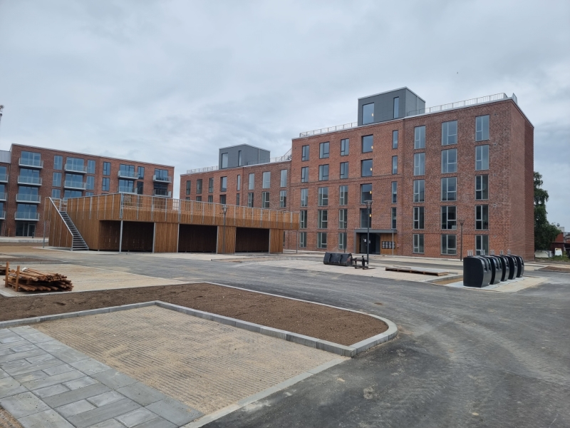 Modern brick apartment buildings with large windows and a wooden rooftop structure in a paved courtyard under a cloudy sky.