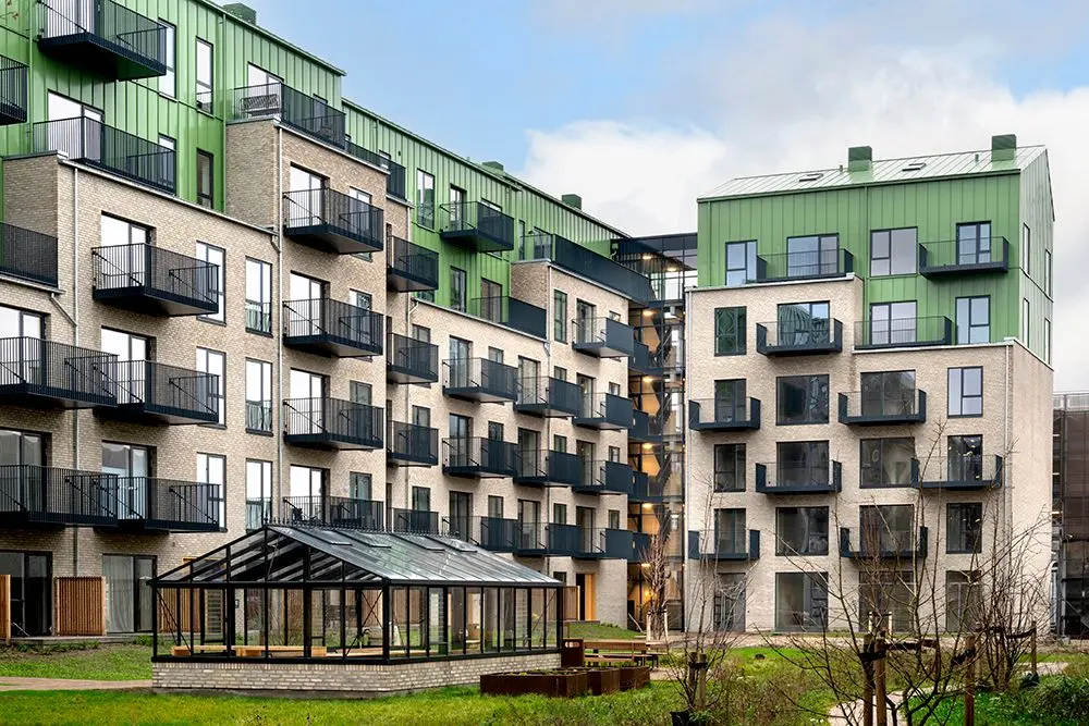 Modern residential buildings with beige brick lower stories and green upper stories, featuring black balconies and a glass greenhouse structure on a lawn.