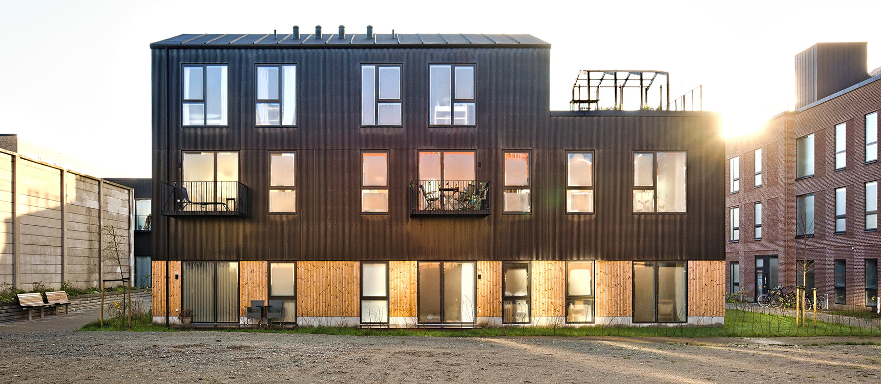 Modern two-story residential building with black upper facade, wooden lower panels, small balconies, and large windows at sunset.