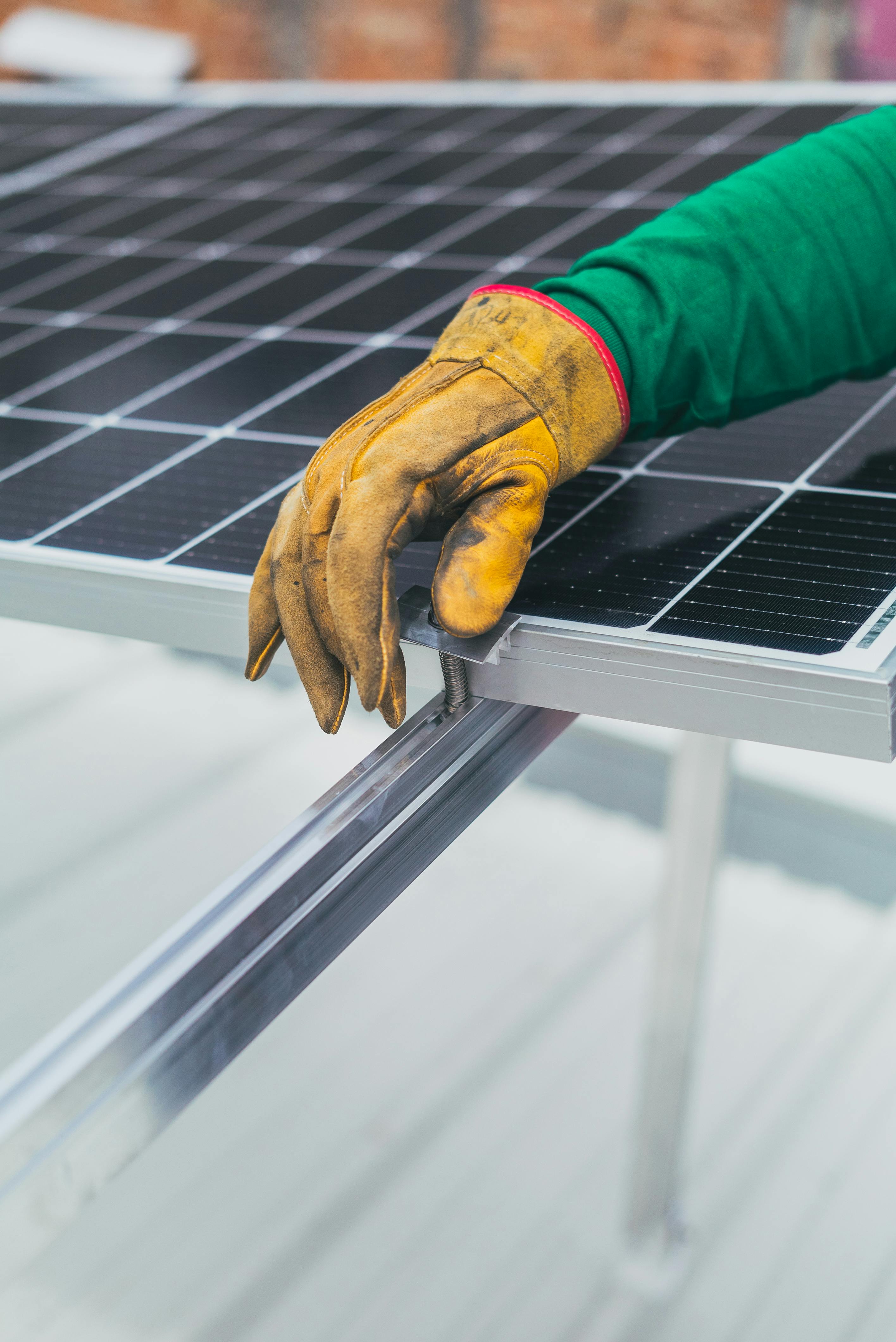Hand wearing a yellow work glove adjusting a clamp on a solar panel frame.