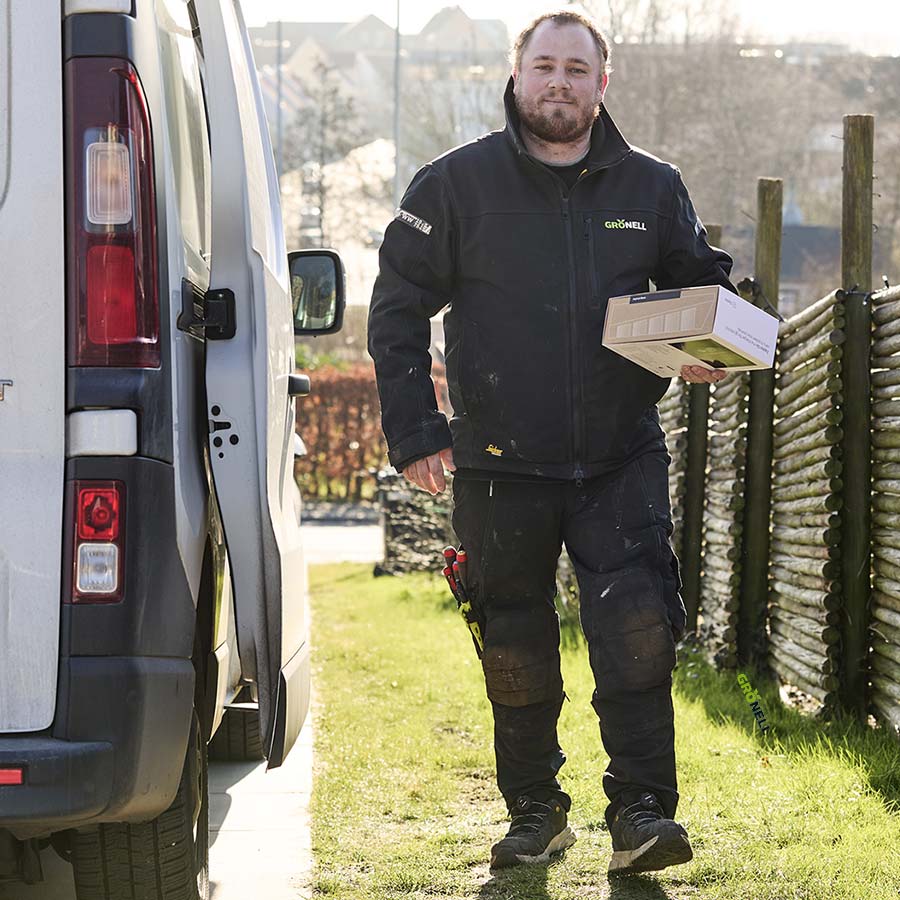 Man wearing a black Grönell jacket carrying a box walking outside beside a white van and wooden fence on a sunny day.