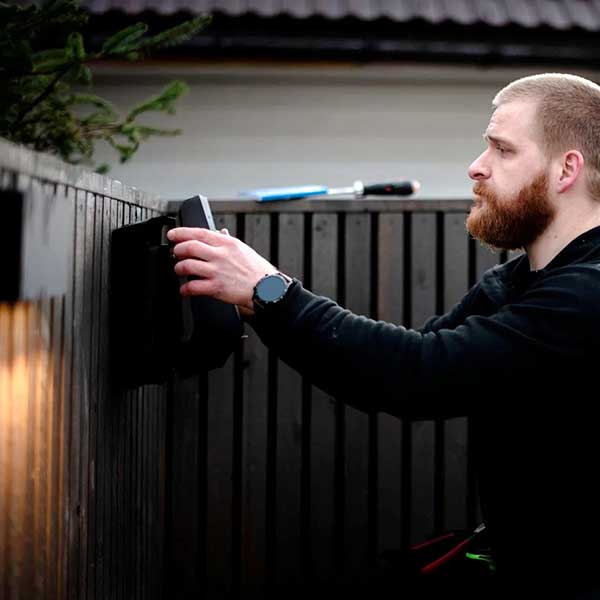 Man with a beard installing or adjusting a black EV charger on a dark wooden fence outdoors.