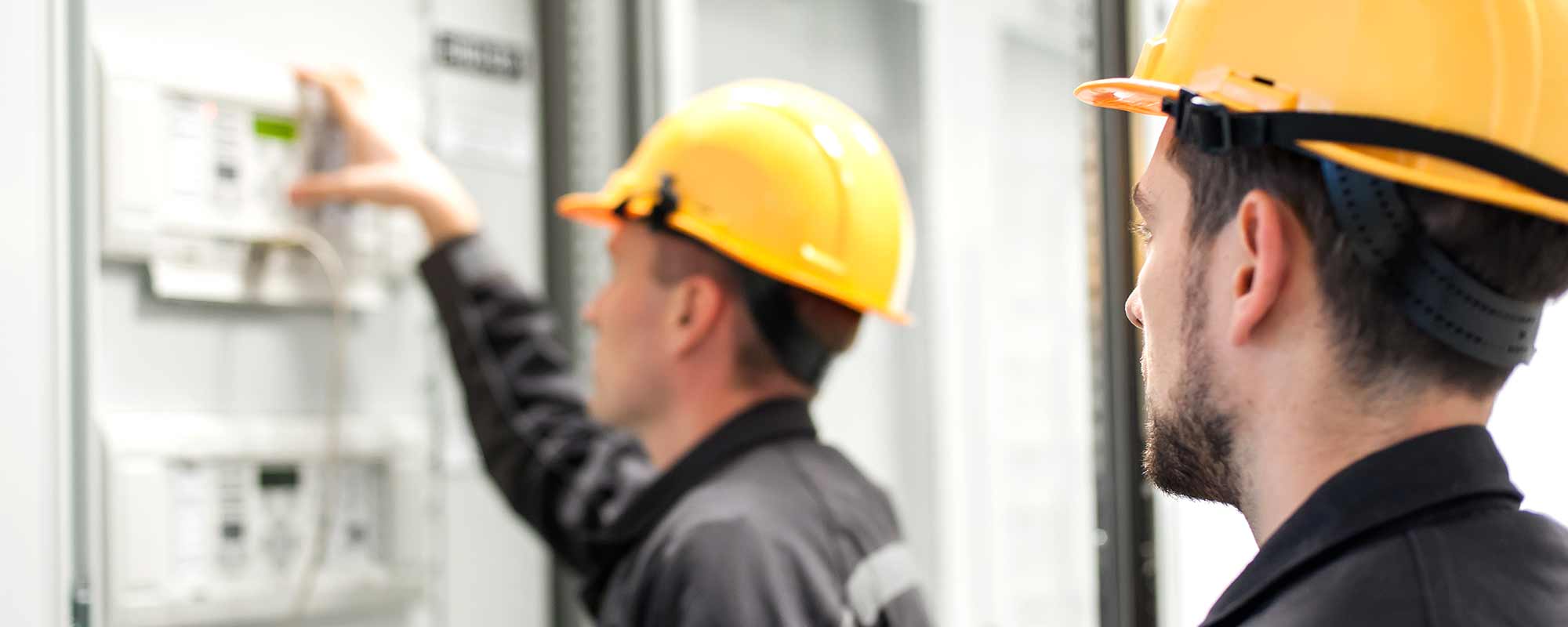 Two male electricians wearing yellow hard hats and black work uniforms inspecting an electrical control panel.