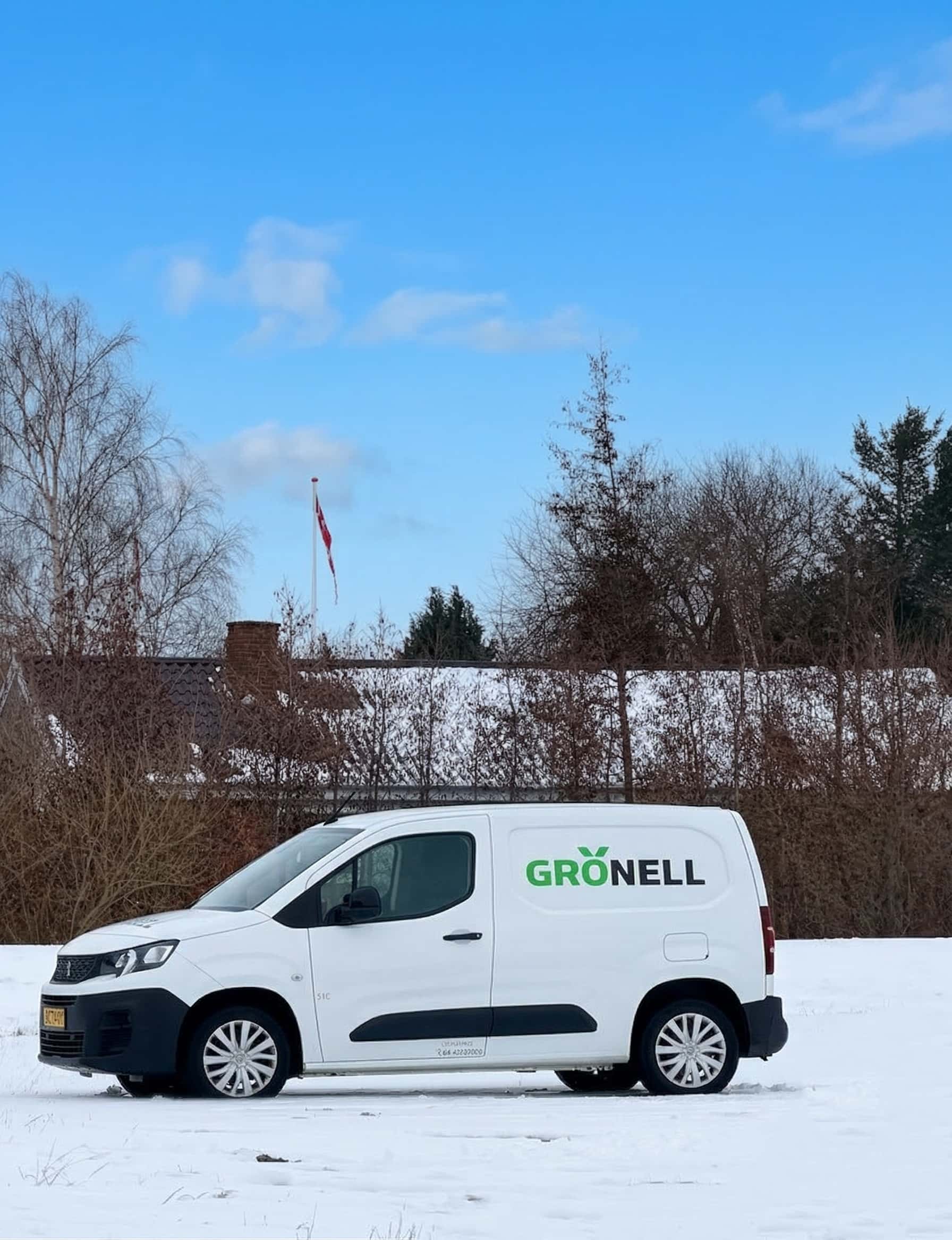 White delivery van with the logo 'GRÖNELL' parked on a snow-covered field with leafless trees and a house in the background under a blue sky.