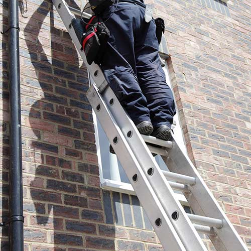Worker in navy uniform climbing an aluminum ladder against a brick wall near a small window.