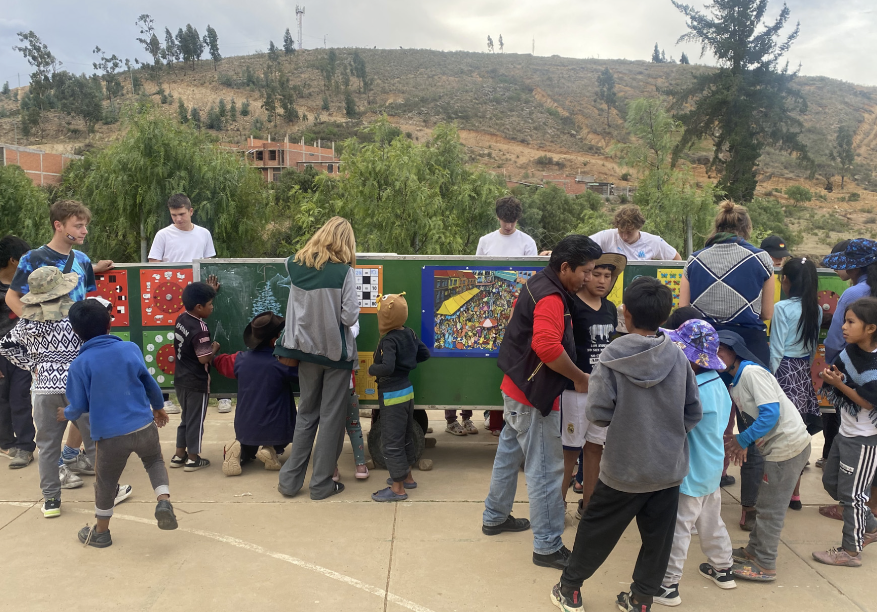 Grupo de niños y adultos interactuando con paneles de actividades educativas al aire libre en una zona rural montañosa.
