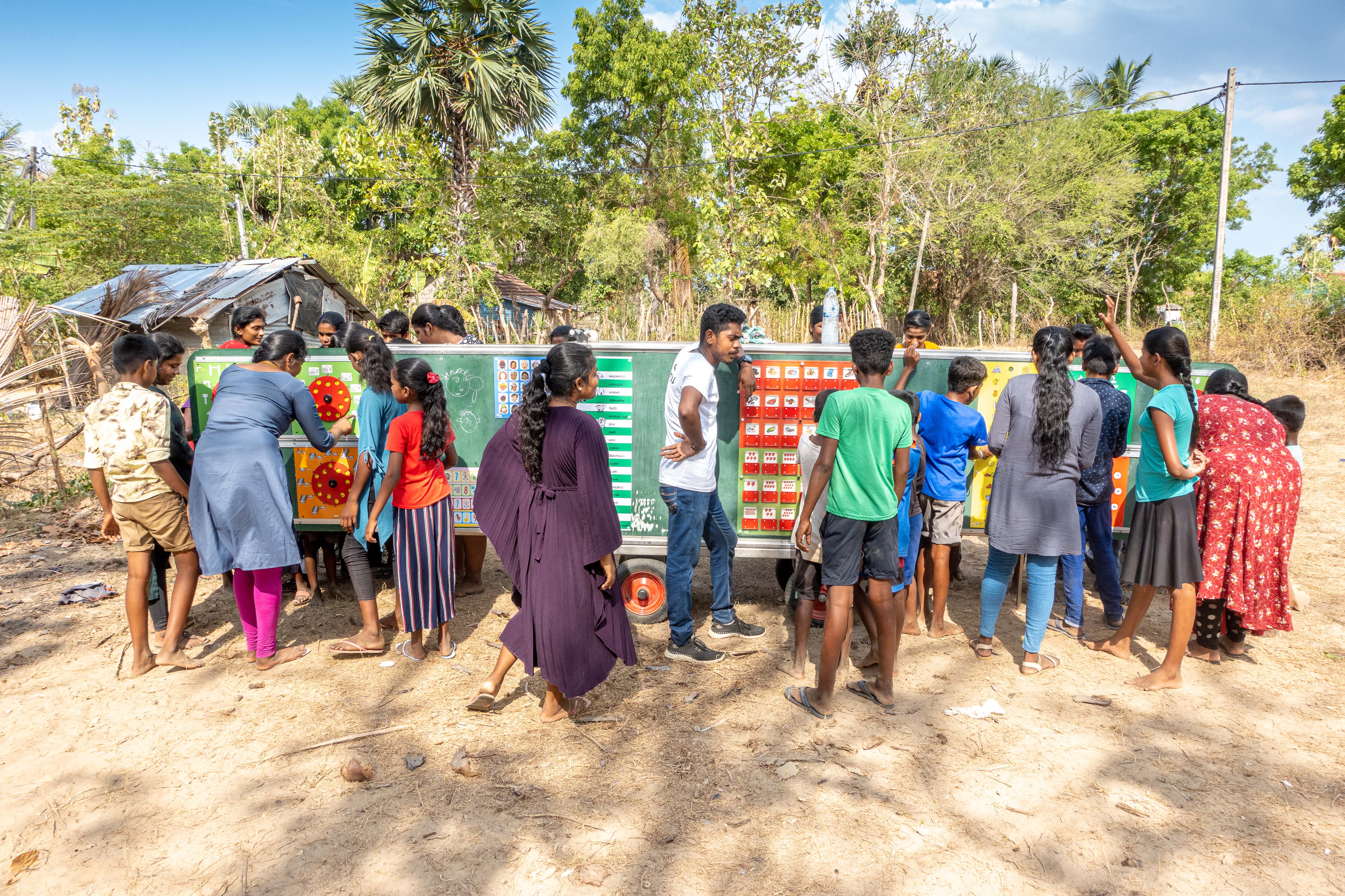 Group of children and adults gathered outdoors around a large mobile educational board with colorful learning materials, Mobile School