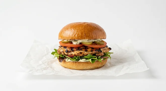 LEON Chargrilled Chicken Burger, chargrilled chicken thigh with tomato, gherkins, leaves and tarragon mayo in a sourdough bun on white paper, nutrition and calories