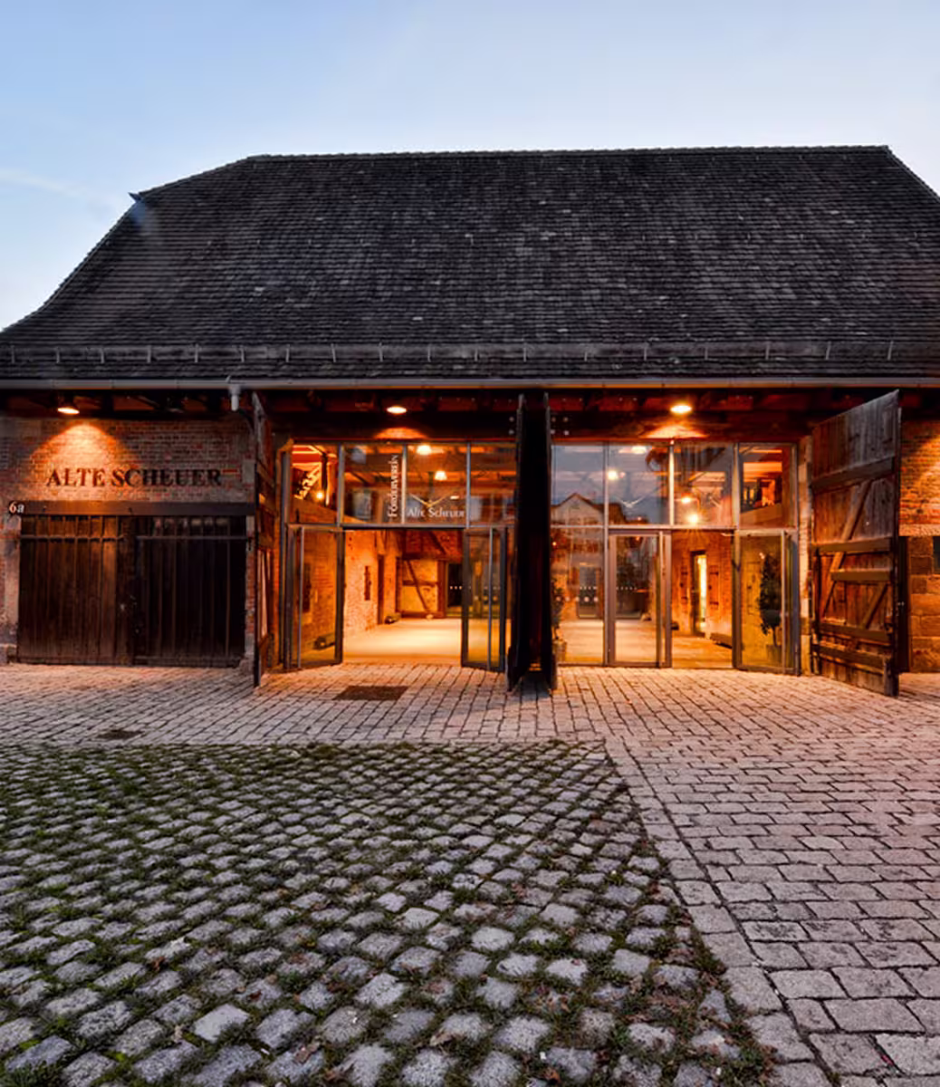 Beleuchtete historische Scheune mit offenem Eingangsbereich und gepflastertem Hof bei Abenddämmerung.