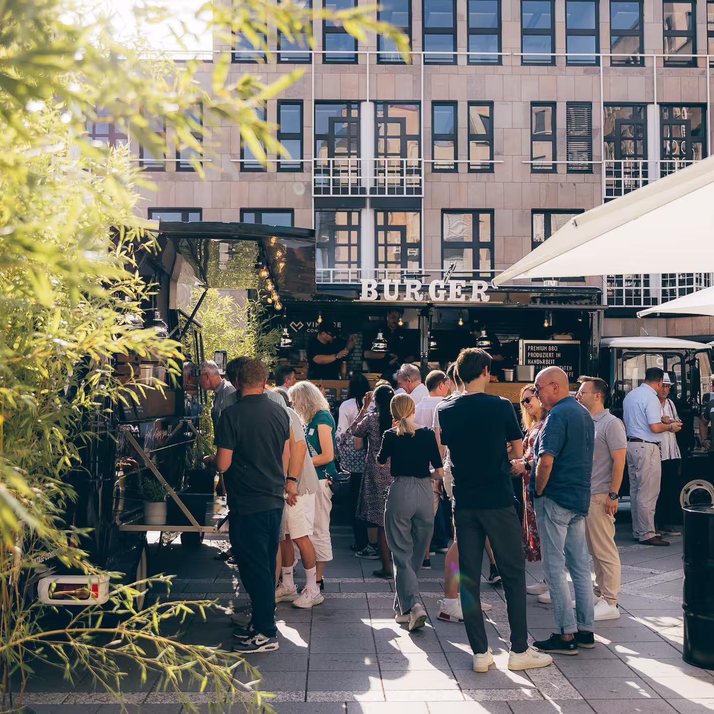 Menschen stehen in einer Schlange vor einem Foodtruck mit der Aufschrift 'BURGER' in einem sonnigen Stadtplatz.