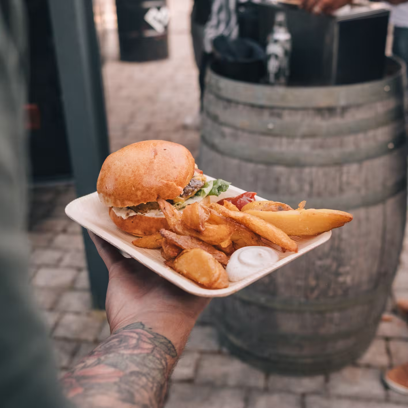 Hand mit Tattoohalted eine Papierschale mit einem Burger, Kartoffelspalten und einem Klecks Mayonnaise.