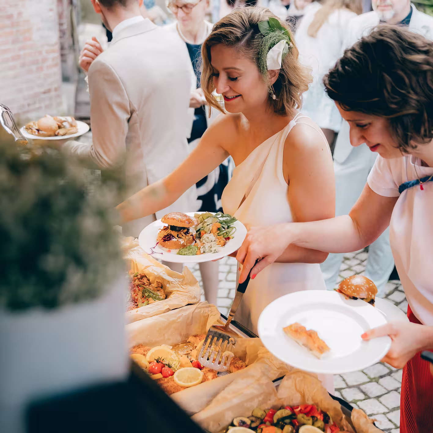 Zwei Frauen nehmen sich Essen von einem Buffet bei einer Feier im Freien, eine hat ein Sandwich und Salat auf dem Teller.