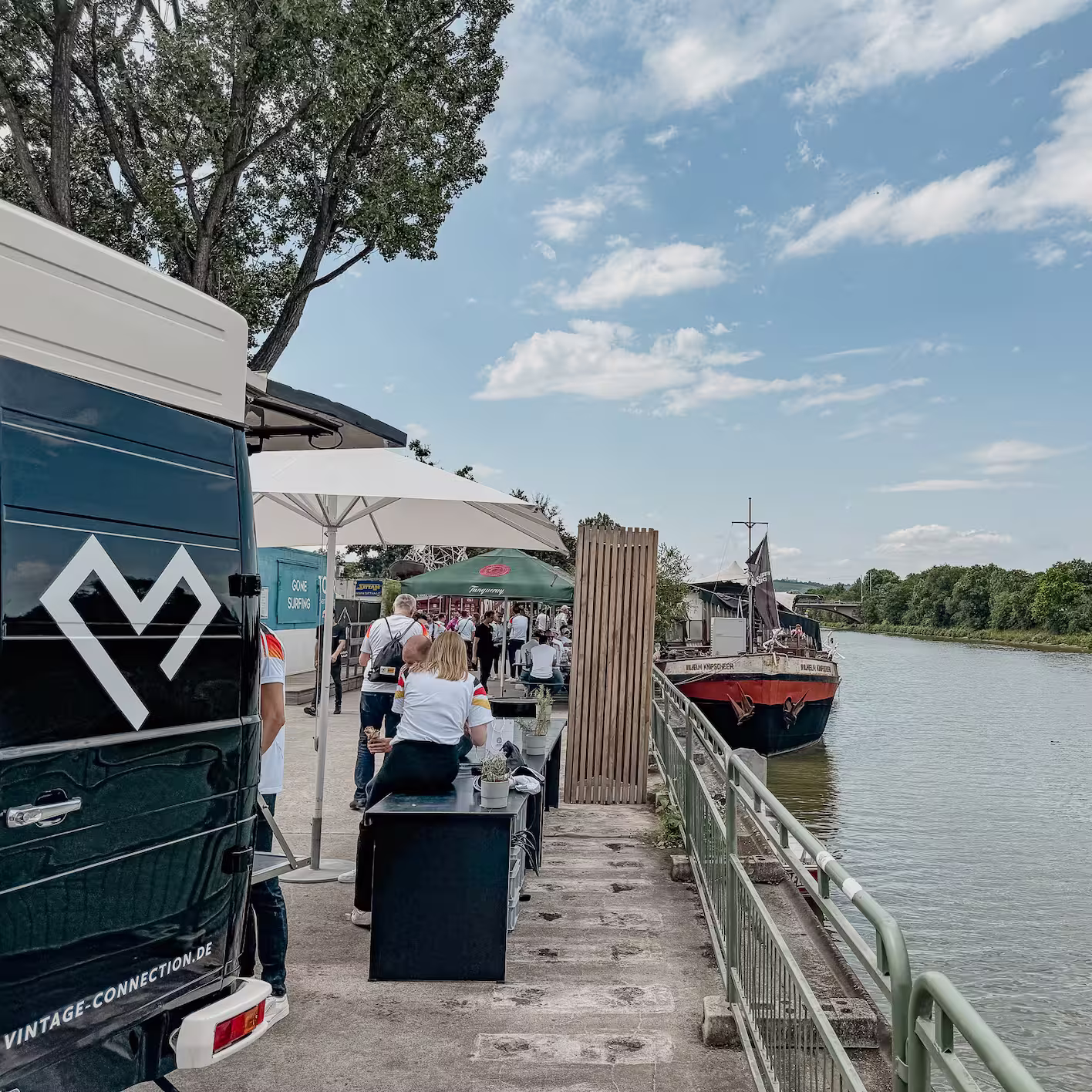 Menschen an einem Flussufer mit Boot und Schirm, daneben ein schwarzer Lieferwagen mit Vintage-Connection-Logo.