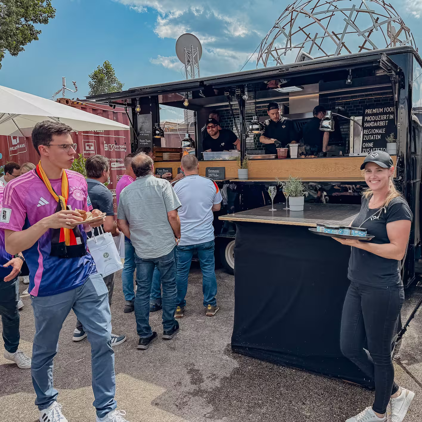 Menschen stehen an einem Foodtruck mit schwarzer Außenwand, während eine Frau mit einem Tablett lächelt und ein junger Mann in Trikot isst.
