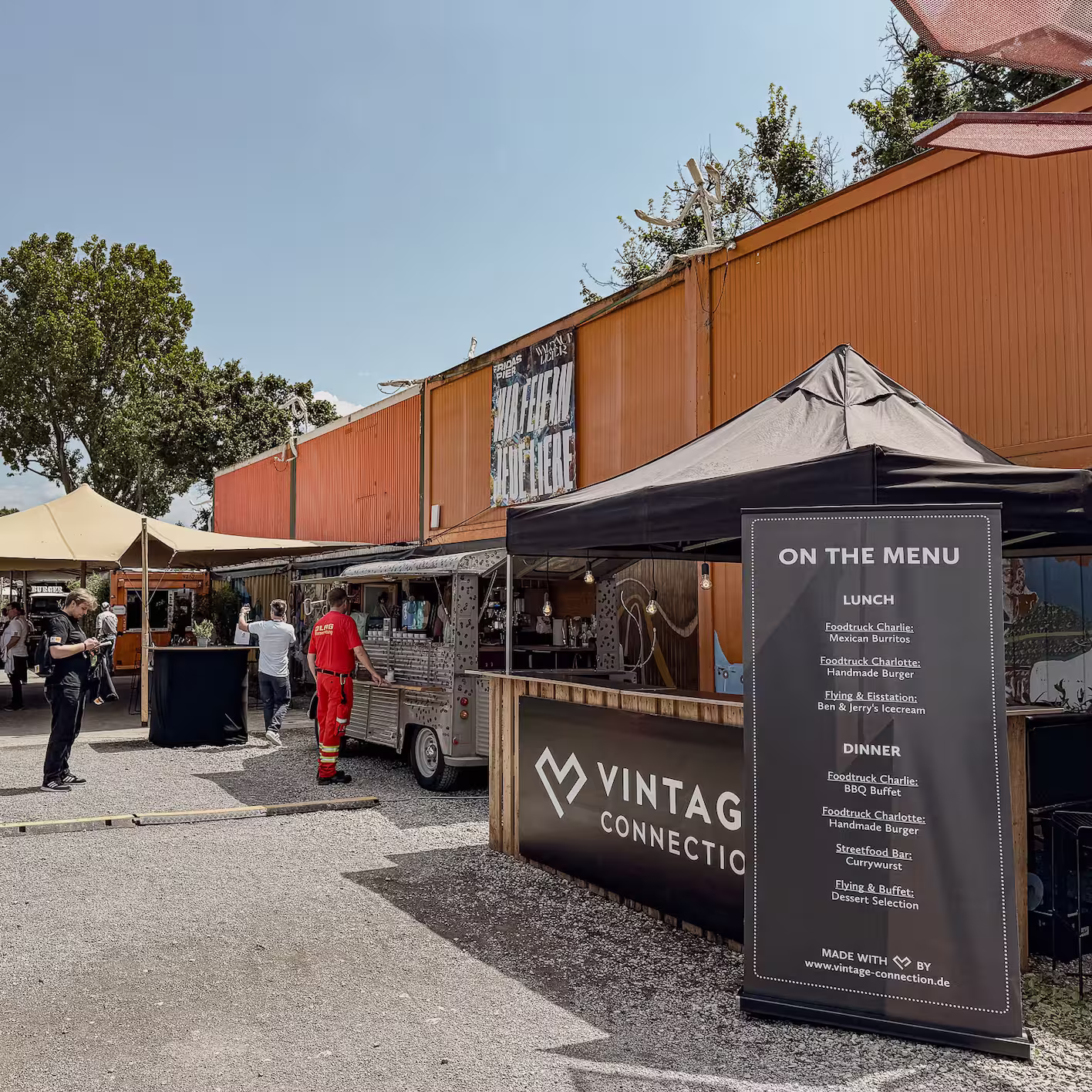 Outdoor Foodtruck-Markt mit mehreren Ständen und einem großen Menü auf Englisch, das Mittag- und Abendessenoptionen auflistet, unter blauem Himmel.
