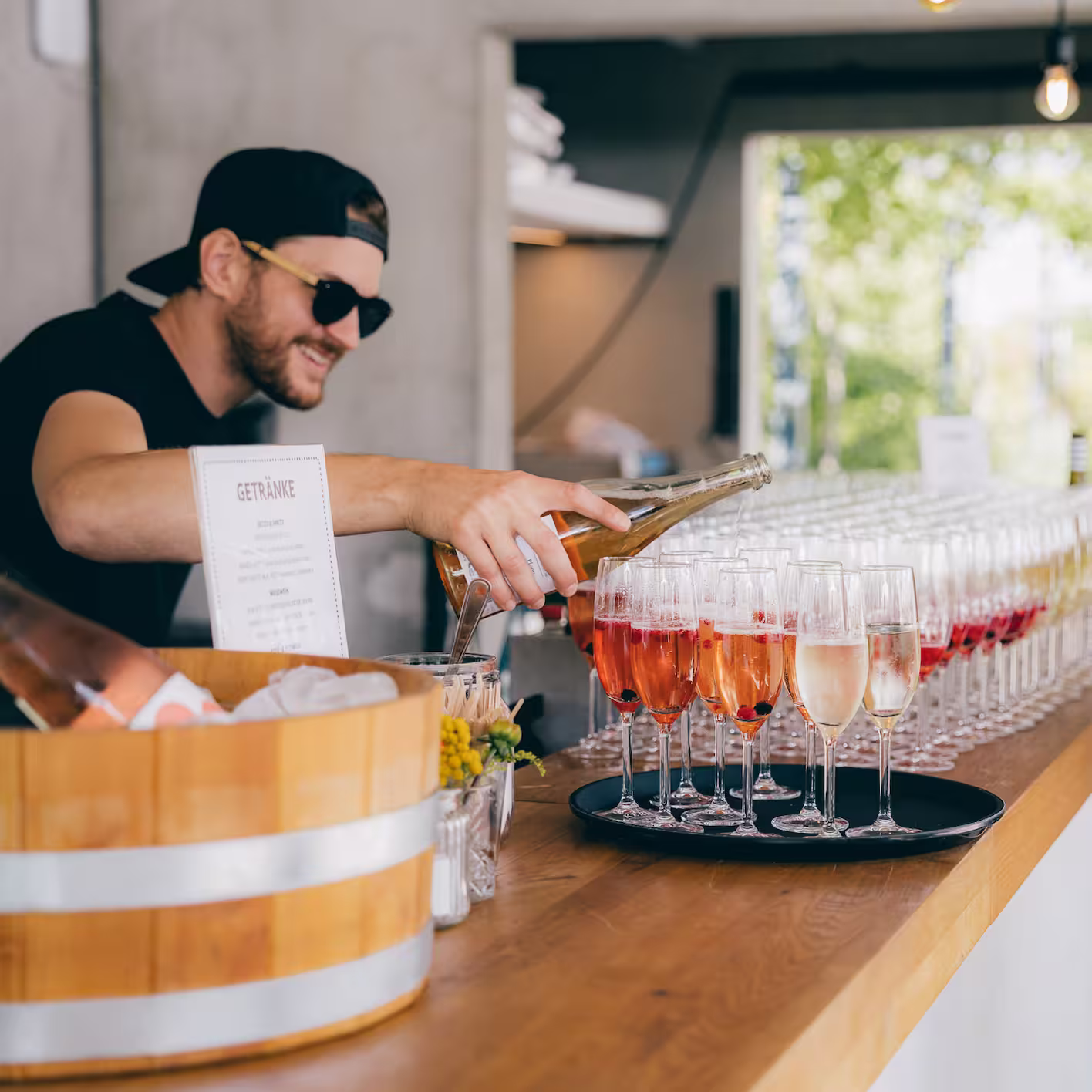 Mann mit schwarzer Kappe und Sonnenbrille gießt Getränke in Reihen von Sektgläsern an einer Bar.