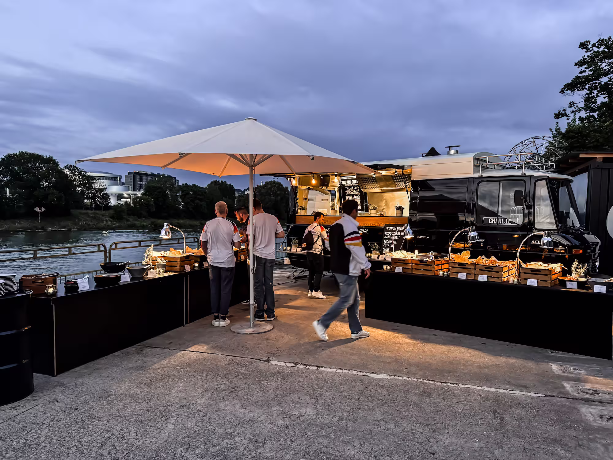 Abendlicher Foodtruck mit beleuchtetem Buffet unter einem weißen Sonnenschirm am Flussufer, umgeben von Menschen.