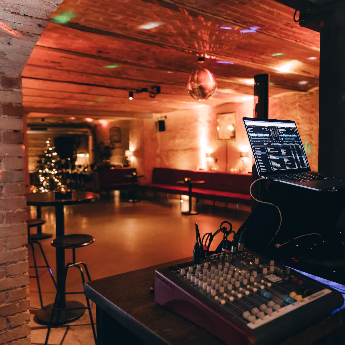 DJ-Setup mit Mischpult und Laptop in einer gemütlich beleuchteten Bar mit Weihnachtsbaum und Discokugel.