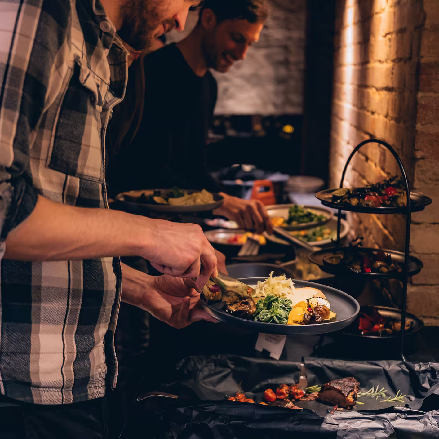 Zwei Männer nehmen Essen von einem Buffet in einem warm beleuchteten Raum mit Backsteinwand.