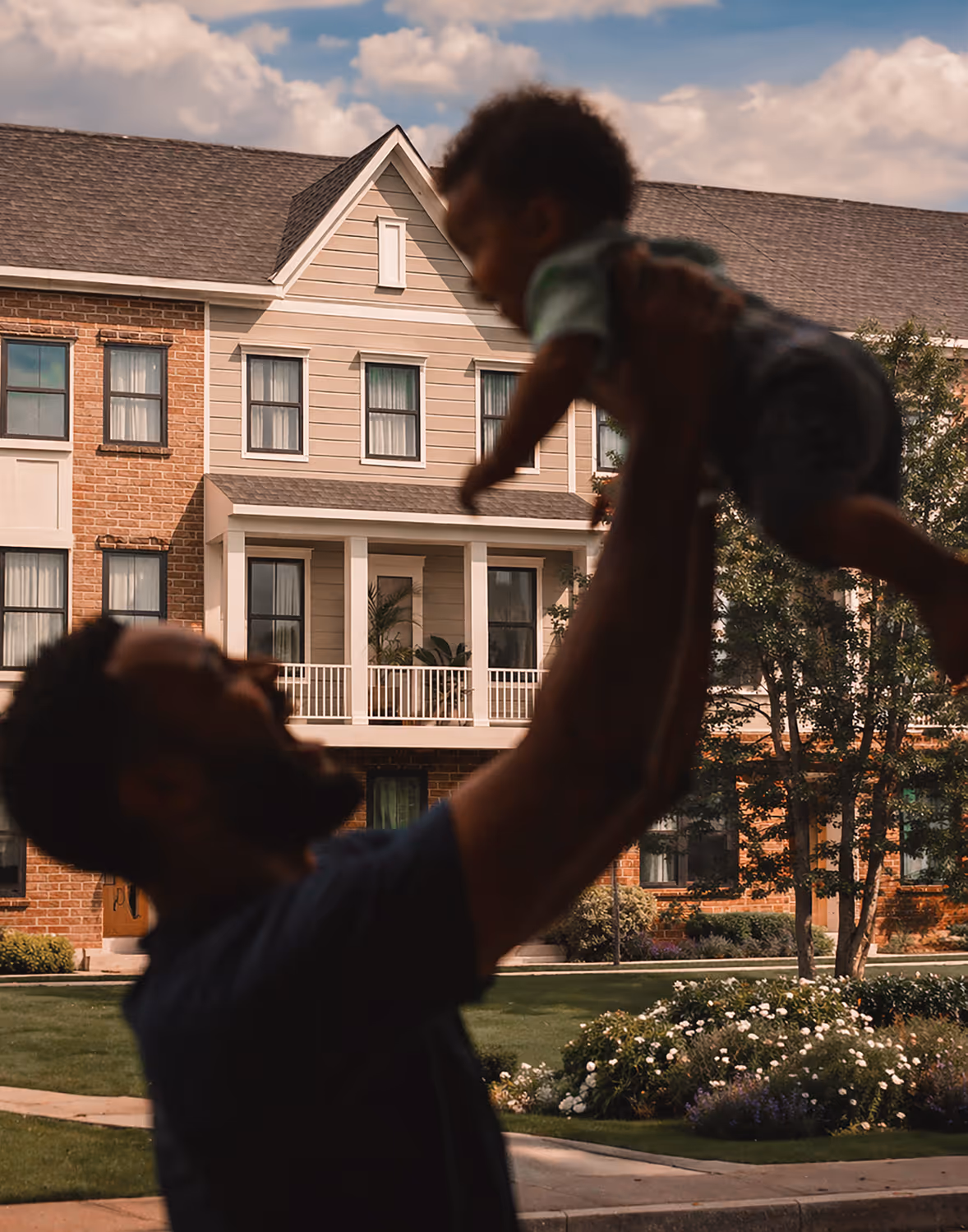 Man lifting smiling baby outdoors in front of multi-story suburban homes with landscaped gardens.