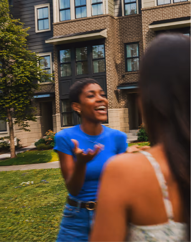 Two women talking and smiling outside in a grassy area with residential buildings in the background.