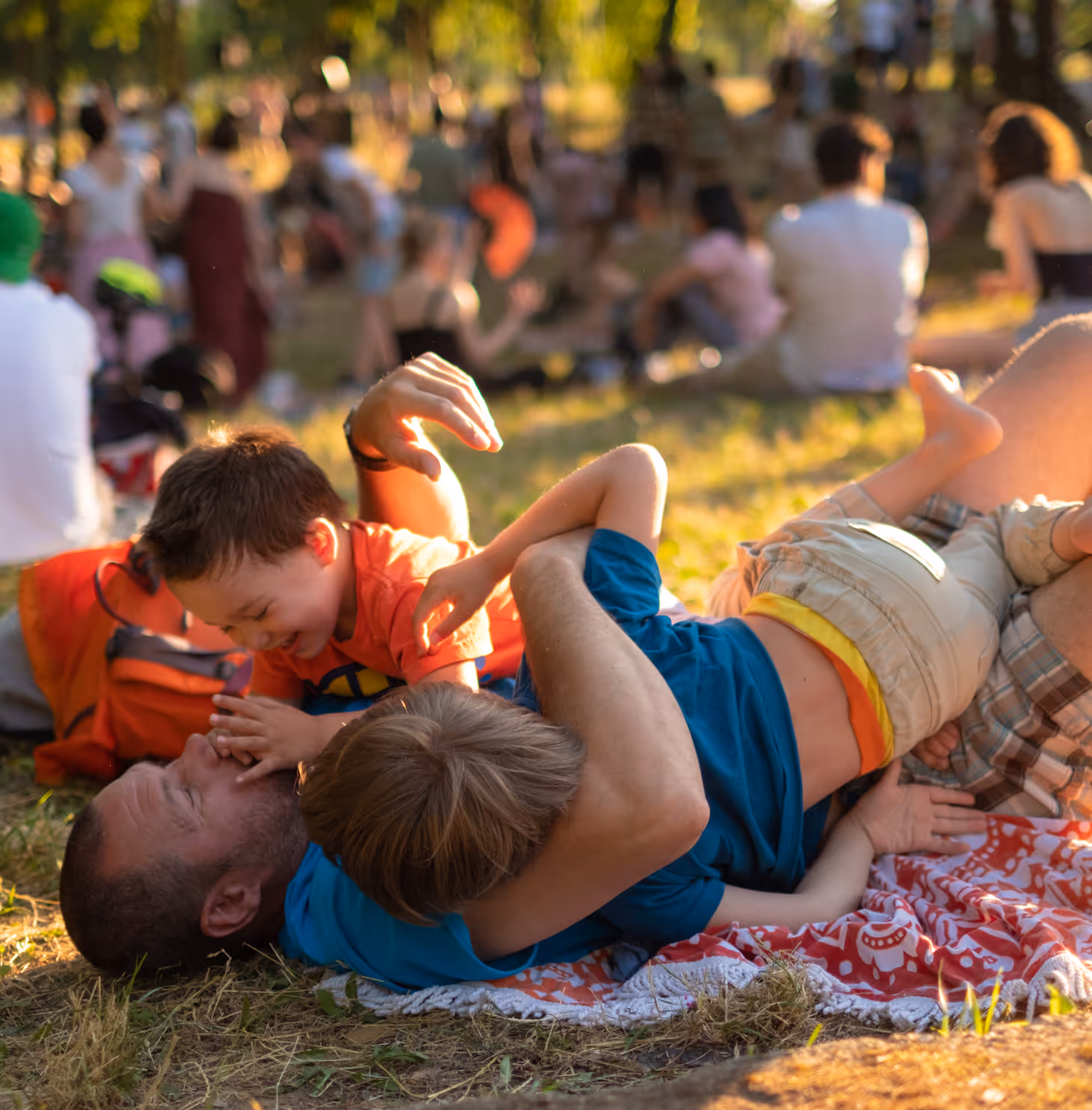 Father playfully lying on grass with two young children during a sunny outdoor gathering.