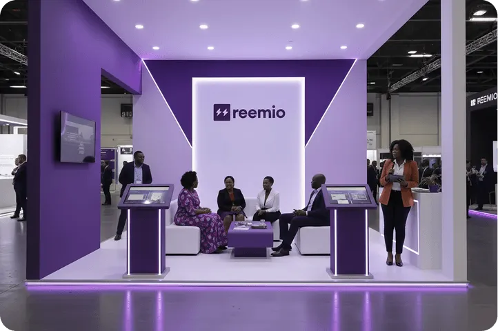 Modern exhibition booth with purple and white design featuring the reemio logo, seating area with four people conversing, and two interactive showcase screens, with a woman standing and presenting on the right side.