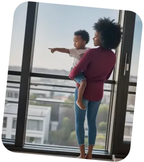 Woman holding a child who is pointing out of a large window overlooking buildings and greenery.