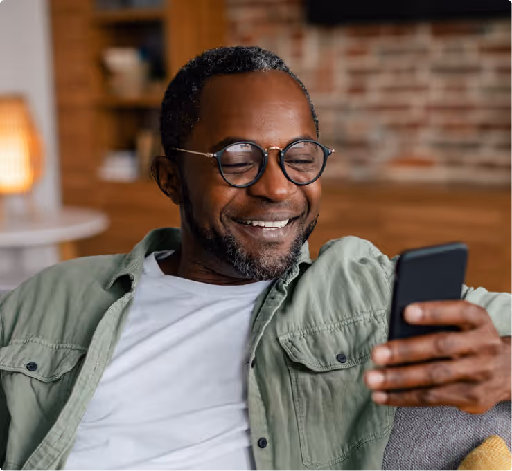 Smiling man with glasses sitting on a couch looking at his smartphone.